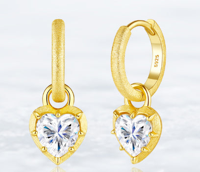 Close-up of a pair of gold hoop earrings featuring heart-shaped crystal pendants on a white background.