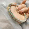 Hand holding a thick silver bangle with intricate colorful enamel scrollwork in shades of green, blue, and gold.