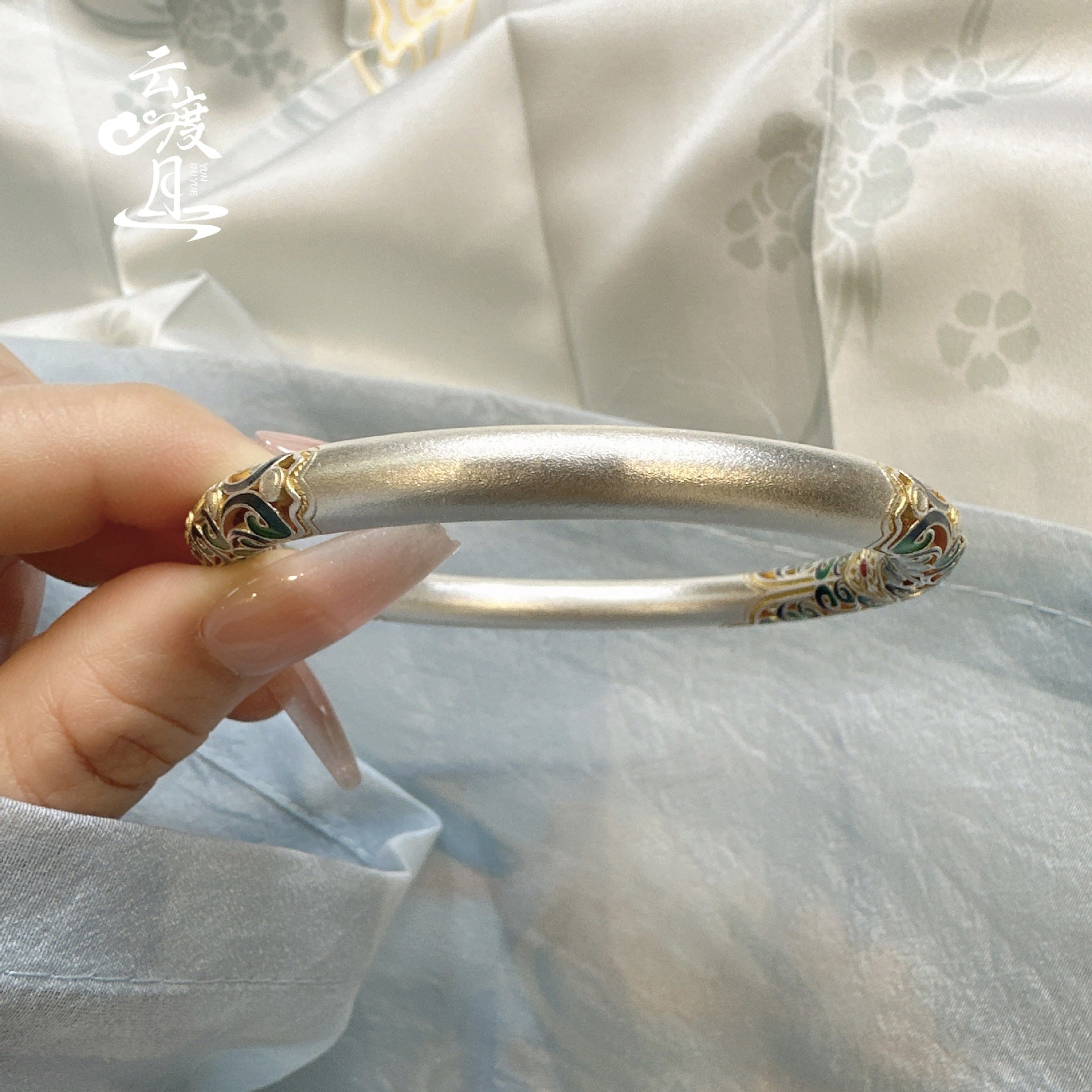 Hand holding a thick silver bangle with intricate colorful enamel scrollwork in shades of green, blue, and gold.