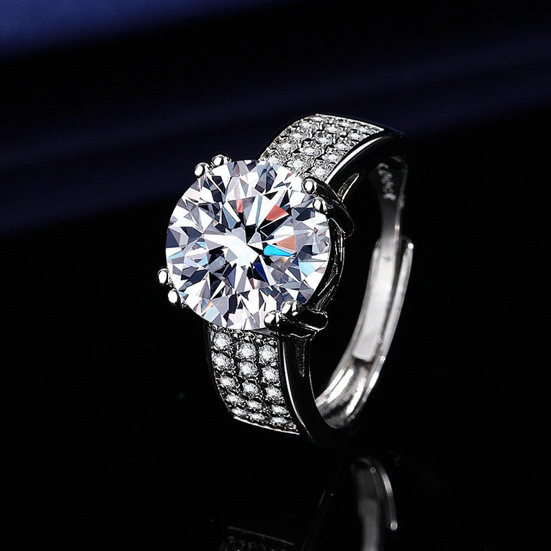 Women's silver ring featuring a large round brilliant-cut center stone and pave-set crystals on a wide band, shown on a dark background.