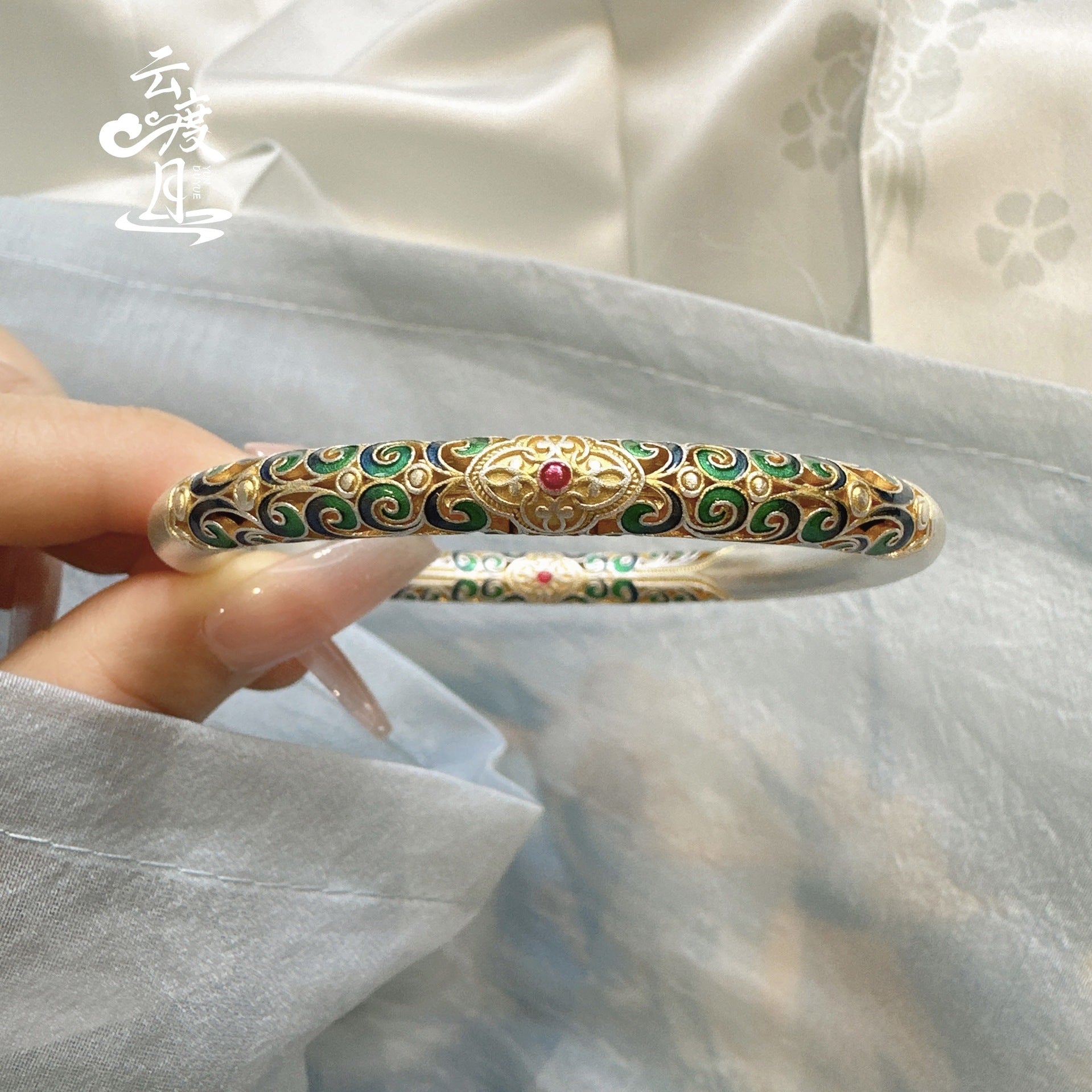 Hand holding a thick silver bangle with intricate colorful enamel scrollwork in shades of green, blue, and gold.