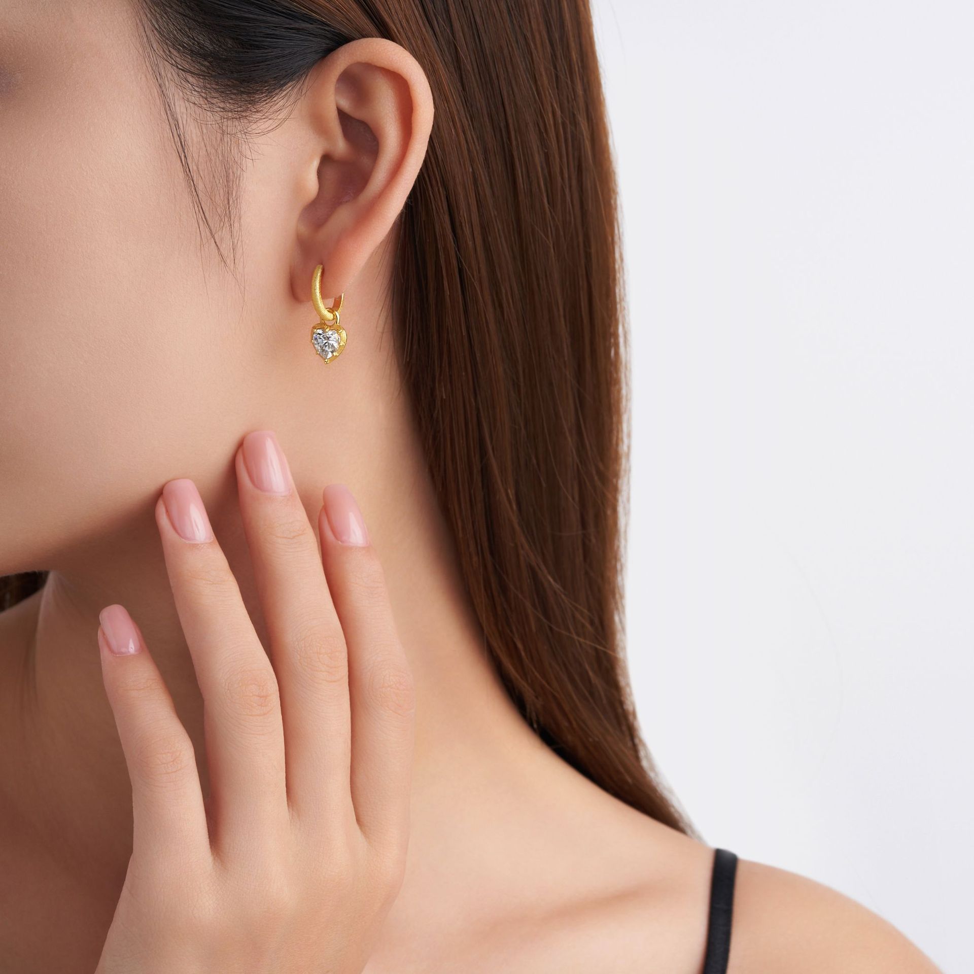 Gold hoop earring with a sparkling heart-shaped crystal pendant, shown worn on a person's ear.