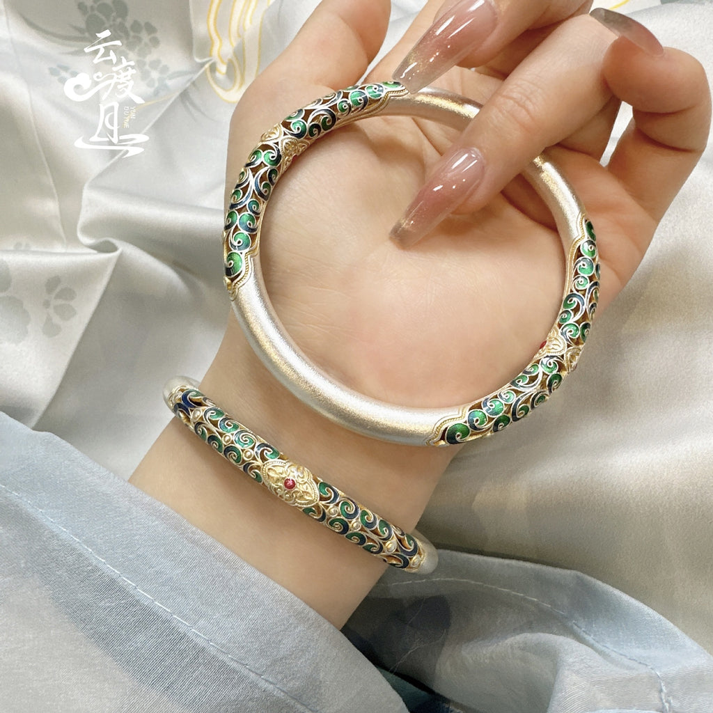 Hand holding a thick silver bangle with intricate colorful enamel scrollwork in shades of green, blue, and gold.