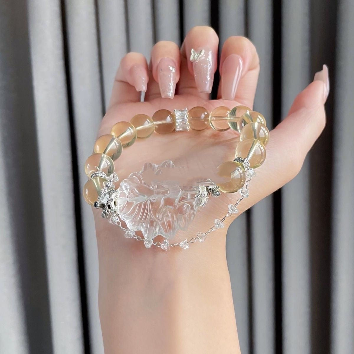 Hand showcasing a yellow beaded bracelet with a large clear carved floral centerpiece and a delicate silver chain accent against a neutral background.