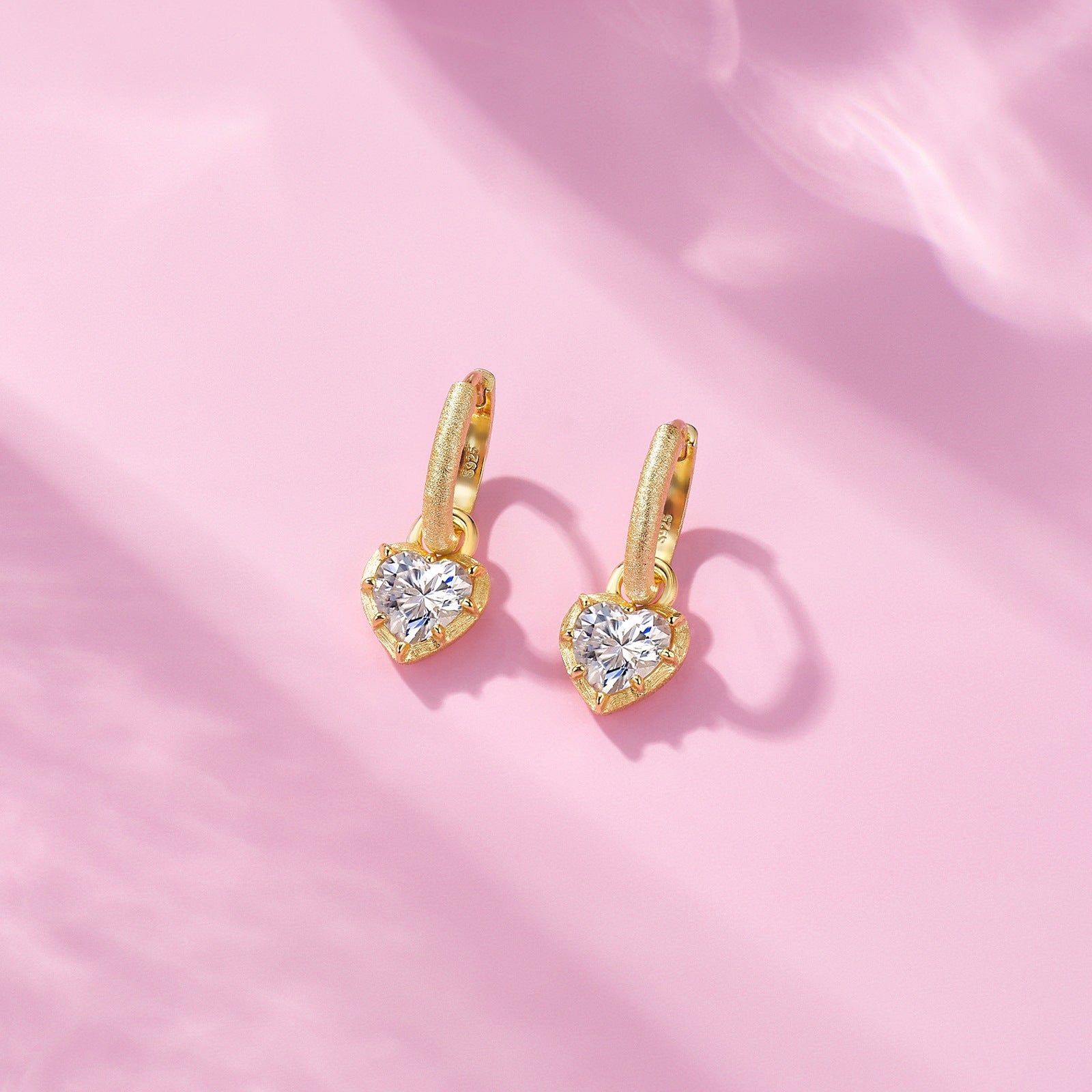 Pair of gold hoop earrings with heart-shaped crystal charms on a soft pink background.