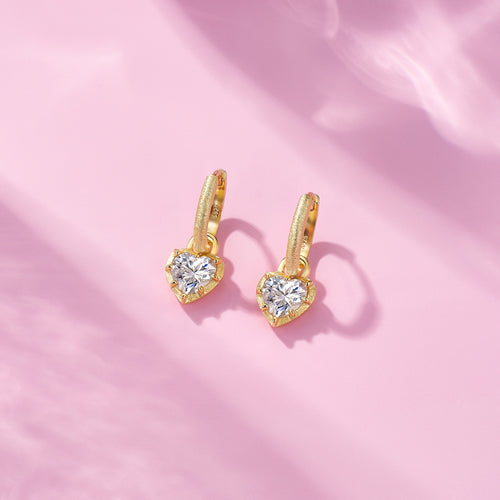 Pair of gold hoop earrings with heart-shaped crystal charms on a soft pink background.