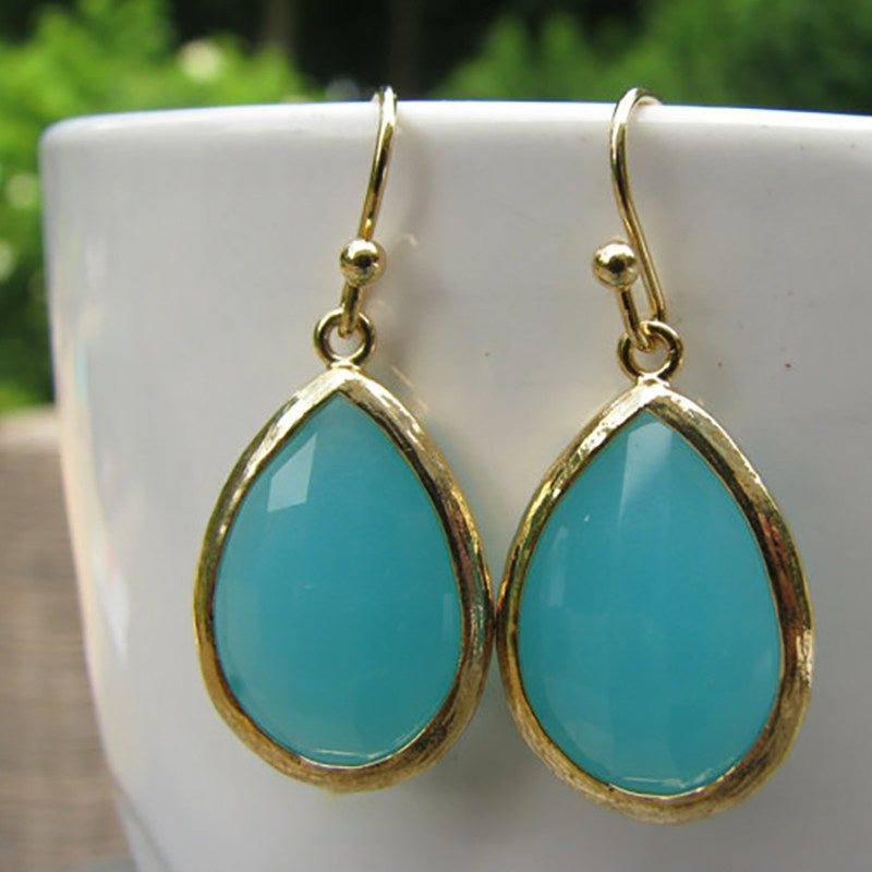 A pair of gold-rimmed teardrop earrings with turquoise blue stones, hanging on the edge of a white ceramic cup.