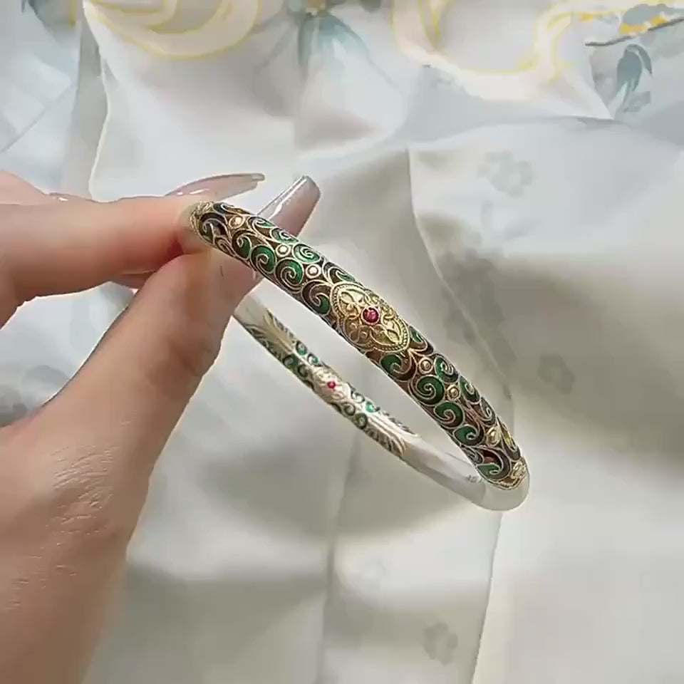 Hand holding a thick silver bangle with intricate colorful enamel scrollwork in shades of green, blue, and gold.